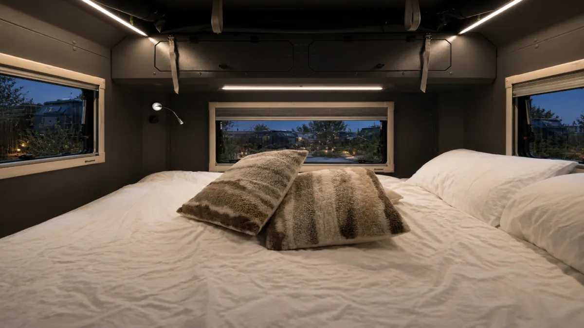 The bed of a caravan made up in white linen, the lit camp visible through three windows